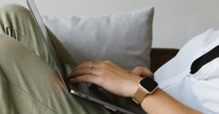 Smart Investments - Side view of crop unrecognizable female freelancer in casual clothes and smartwatch lying on comfortable sofa and working remotely on laptop