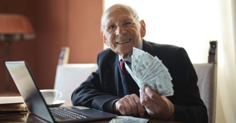 Smart Investments - Happy senior businessman holding money in hand while working on laptop at table
